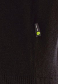 Schwarzer Strickpullover mit geripptem Saum, ausgestattet mit einem kleinen Etikett in Grün und Grau mit Text. Weiche Textur und runder Ausschnitt.