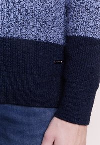 Stickad tröja i blått och marinblått med texturerat tyg. Har ett horisontellt randmönster och en liten logotagg nära fållen.