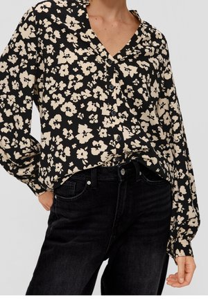 Schwarze Bluse mit floralem Muster in cremfarbenen Tönen, mit V-Ausschnitt, Knopfverschluss und lockeren Ärmeln. Kombiniert mit dunkelblauen Jeans.