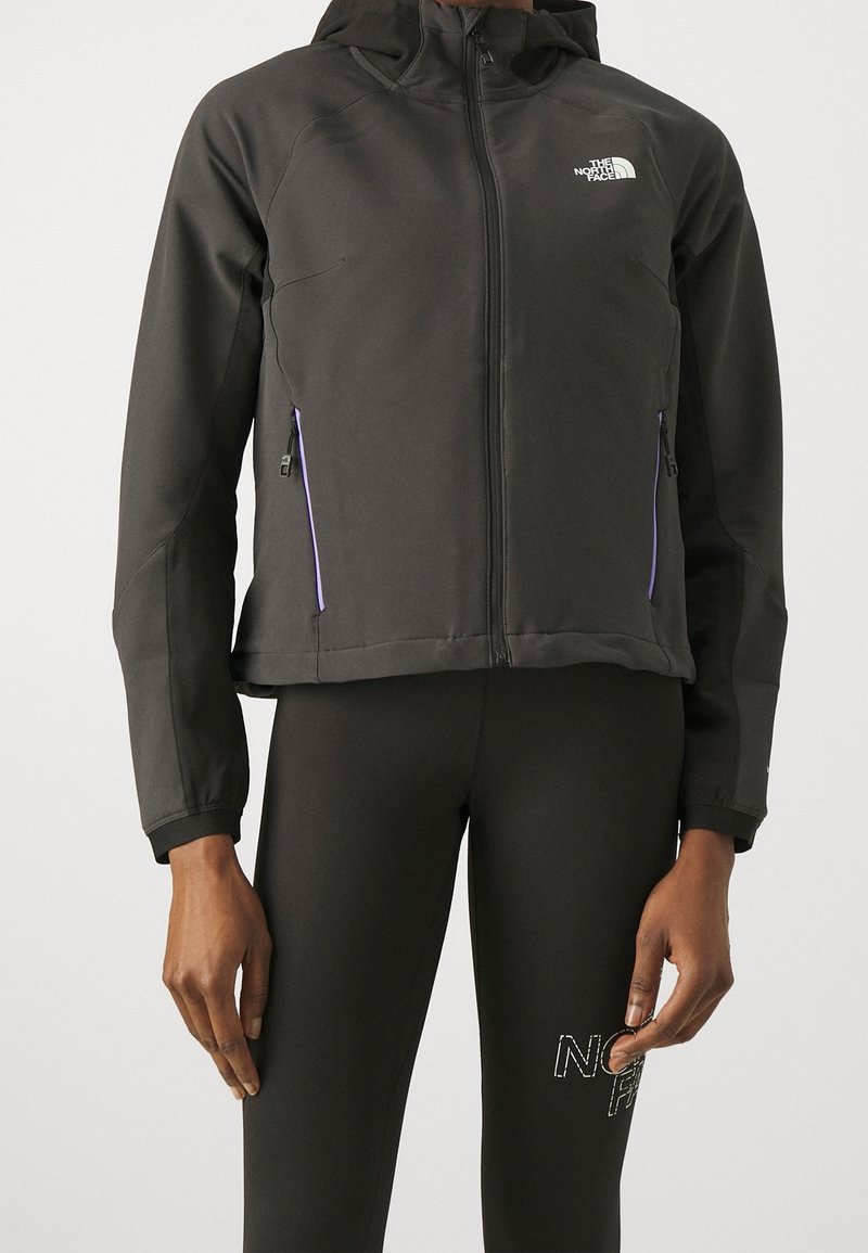 Person som bär en svart jacka med hel dragkedja och huva, med fickor på sidan som också har dragkedja, kombinerat med svarta leggings med en vit logotyp på låret.