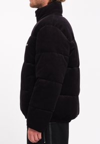 Schwarze Corduroy-Pufferjacke mit hohem Kragen, vertikaler Rippstruktur und elastischem Saum. Die Seitenansicht zeigt die leichte Konstruktion.