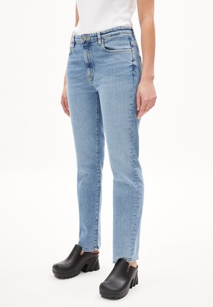 Frau, die hellblaue gerade geschnittene Jeans und schwarze klobige Clogs mit Absatz trägt, steht vor weißem Hintergrund.