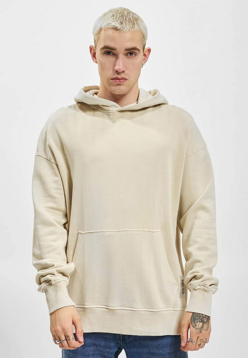 Sweat-shirt à capuche beige avec une poche avant, des épaules tombantes et des poignets côtelés, fabriqué à partir d'un tissu doux, présentant une texture subtile.