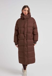 Bruine pufferjas met hoge kraag, lange lengte en gequilt ontwerp. Voorzien van ritszakken en brede mouwen voor extra comfort.