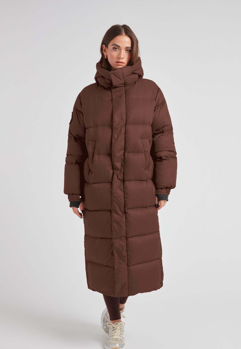 Bruine pufferjas met hoge kraag, lange lengte en gequilt ontwerp. Voorzien van ritszakken en brede mouwen voor extra comfort.