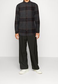 Dunkelgraues Flanellhemd mit einem Karomuster in Braun- und Rottönen. Langärmlig, mit Knopfleiste, kombiniert mit schwarzer Hose und weißen Turnschuhen.