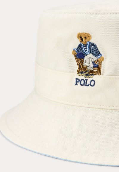 Chapeau bob crème avec un ours brodé portant un blazer bleu et un pantalon blanc, assis sur une chaise au-dessus du mot « POLO ».
