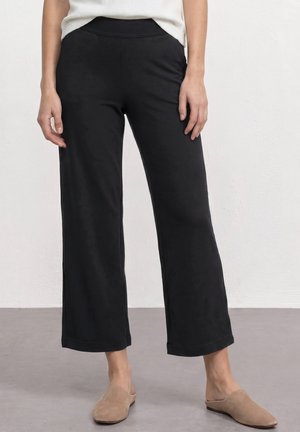 Femme portant un pantalon noir à jambes larges, un haut blanc et des chaussures beige sans lacets, debout devant un mur et un sol clairs et unis.