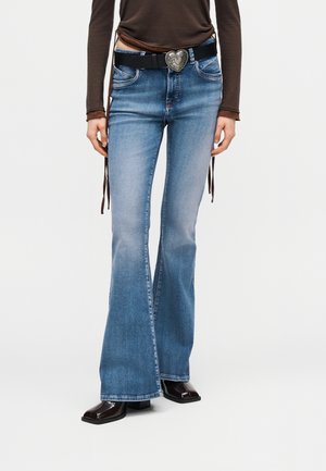 Άτομο που φοράει μπλε καμπάνα τζιν, μαύρη ζώνη με αγκράφα σε σχήμα καρδιάς, καφέ μακρυμάνικη μπλούζα και σκούρα παπούτσια με τετράγωνη μύτη.