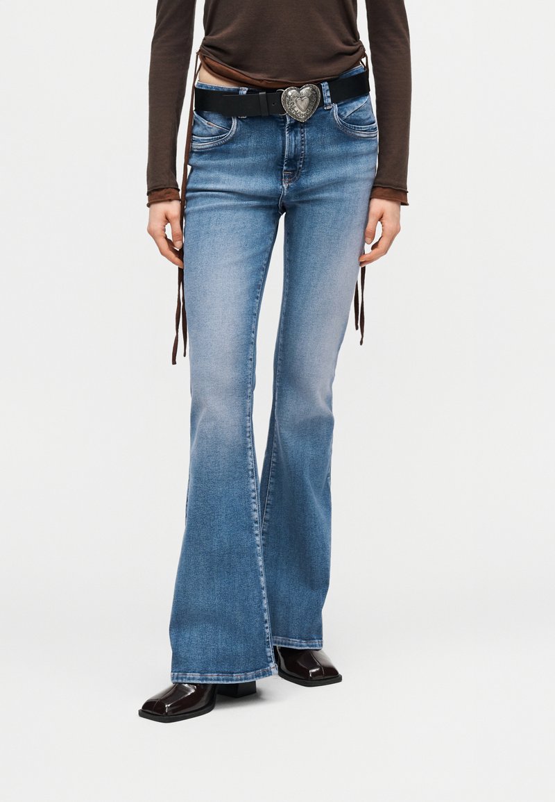 Άτομο που φοράει μπλε καμπάνα τζιν, μαύρη ζώνη με αγκράφα σε σχήμα καρδιάς, καφέ μακρυμάνικη μπλούζα και σκούρα παπούτσια με τετράγωνη μύτη.