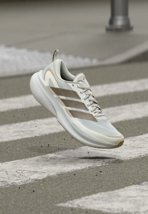 Pantof de alergare Adidas alb și bej, în aer deasupra unui drum asfaltat gri cu linii albe de trecere pentru pietoni.