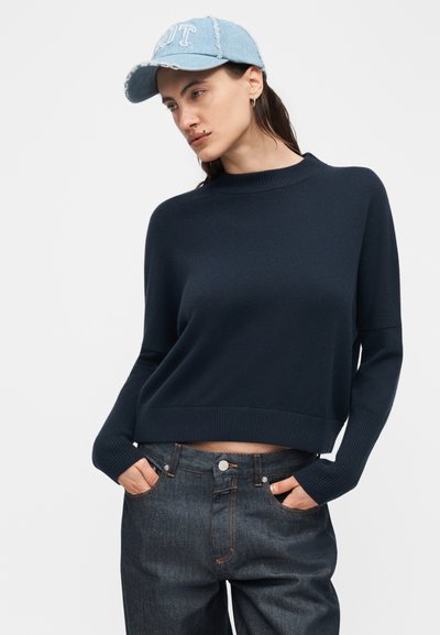Femme portant un pull bleu marine, un jean en denim foncé avec les mains dans les poches, et une casquette en denim bleu clair avec un lettrage blanc sur un fond uni.