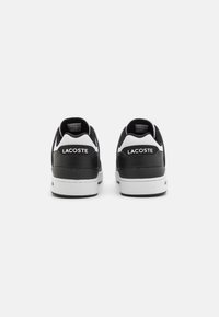 Zapatillas en blanco y negro con una parte superior de cuero suave, acentos texturizados y un tacón pronunciado, con el logo de "Lacoste" en blanco.