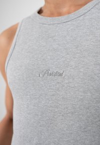 Gros plan d'une personne portant un débardeur gris clair côtelé avec une broderie cursive discrète sur la poitrine.