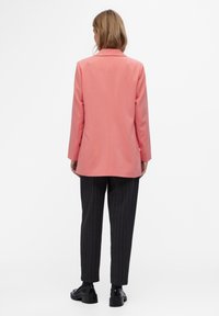 Coral blazer with a structured design, long sleeves, and a single back vent, paired with black pinstriped trousers and shiny black shoes.