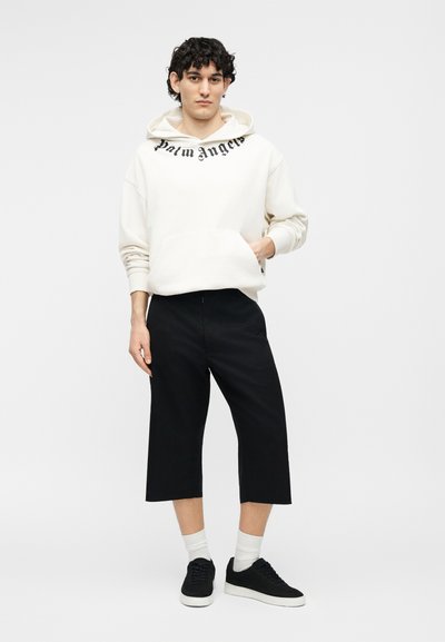 Jeune homme portant un sweat à capuche blanc Palm Angels, un pantalon large cropped noir, des chaussettes blanches et des baskets noires, debout devant un fond blanc uni.