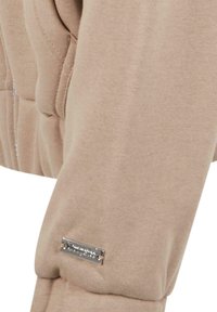 Détail d'un sweat beige présentant une texture douce, une fermeture éclair et un accent en logo argenté sur le bras gauche.