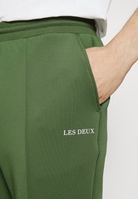Groene joggers gemaakt van gladde stof, met een zijzak en de tekst "LES DEUX" in het wit gedrukt op het been.