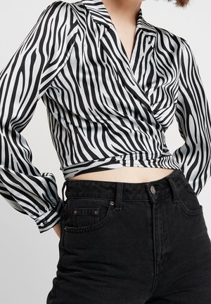 Nainen, jolla on yllään mustavalkoraidallinen seepra-kuvioinen pitkähiha wrap-paita ja korkeavyötäröiset mustat farkut vaaleaa taustaa vasten.