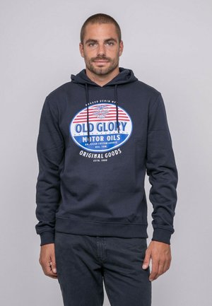 Homme aux cheveux courts et barbe portant un sweat à capuche bleu marine avec le logo "Old Glory Motor Oils", debout devant un fond gris.