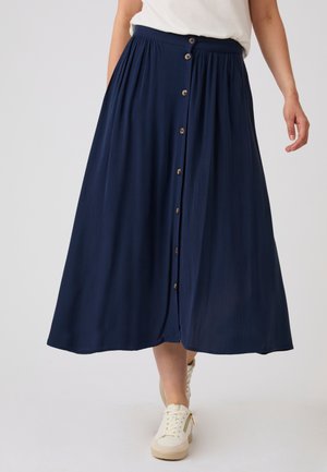 Mujer con una falda midi azul marino con botones delanteros combinada con zapatillas blancas y una camiseta blanca metida por dentro.