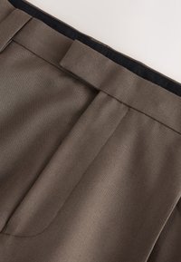 Close-up of brown fabric pants waistband with belt loop and discreet side pocket seam detail.