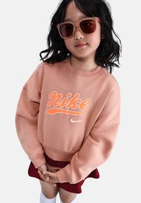 Sudadera rosa de Nike con un gran logo naranja, mangas largas y puños acanalados, combinada con una falda roja y calcetas blancas hasta la rodilla.