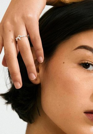 Silver ring with a bow design on a finger, contrasted against smooth skin with natural nails and short dark hair.