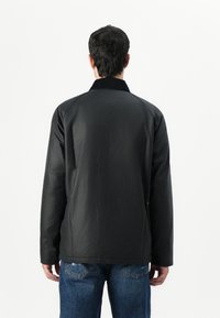 Veste noire à col avec une texture lisse, dotée de manches longues et d'une coupe légèrement ample, associée à un jean en denim bleu.
