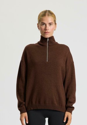 Kobieta z blond włosami, ubrana w brązowy sweter z zamkiem błyskawicznym i czarne spodnie, stojąca na tle jasnego, jednolitego tła.