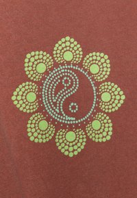 Rostfärgad tyg med ett cirkulärt mönster av gröna och blå prickar, som bildar ett blommönster och ett centralt yin-yang-symbol.