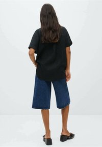 Maglietta nera a maniche corte, dal taglio morbido, con orlo arrotondato. Abbinata a culotte in denim blu lunghe fino al ginocchio. Indossata con sandali neri con suola spessa.
