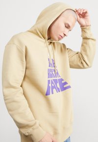 Sudadera marrón con un gráfico de logo púrpura en la parte delantera. Tiene una capucha con cordón, puños de canalé y un ajuste relajado.