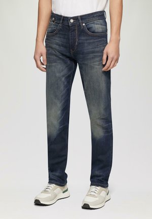 Donkerblauwe jeans met een vervaagde wash, vijf-pocket ontwerp, metalen knoopsluiting en contrasterende oranje stiksels. Draag met lichte sneakers.