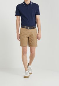 Polo shirt marine avec liseré blanc, shorts beiges et ceinture à motifs, associé à des baskets blanches chunky avec des accents beige.