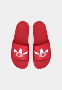 Punaiset kumiset liukusandaalit, joissa on leveä hihna ja valkoinen trefoil-logo, jossa on kolme vaakasuoraa raitaa. Sileä pinta ja pyöristetty muoto.