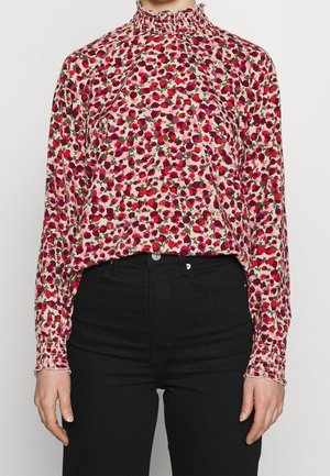 Kvinde iført en lyserød bluse med lange ærmer med rødt og lilla blomsterprint og flæset høj hals, kombineret med sort højtaljede bukser.