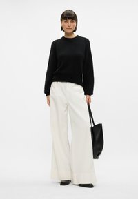 Black knit sweater with long sleeves, paired with white wide-leg trousers and black pointed-toe shoes, holding a black tote bag.