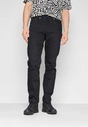 Jeans en denim noir avec une coupe slim, design à cinq poches et quincaillerie métallique visible. Portés avec des chaussures de sport noires et une chemise à motifs.