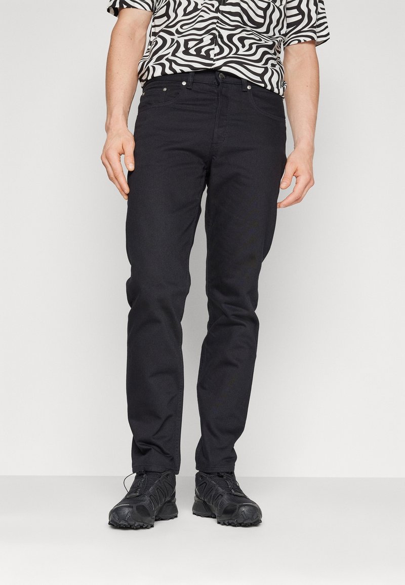 Jeans de mezclilla negra con un corte ajustado, diseño de cinco bolsillos y herrajes metálicos visibles. Llevados con zapatillas deportivas negras y una camisa con estampado.