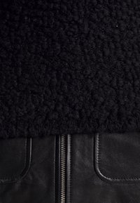 Tissu en peau lainée noire au-dessus de cuir noir avec une fermeture éclair argentée centrale et deux poches en cuir cousues en bas.