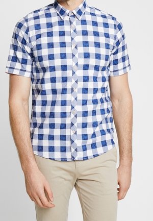 Homme portant une chemise à manches courtes à carreaux bleus et blancs, boutonnée, associée à un pantalon beige clair sur fond blanc uni.
