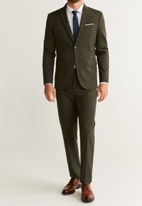 Completo verde oliva con giacca a due bottoni, revers a punta e pantaloni abbinati. Camicia bianca e cravatta blu navy, completati da scarpe marroni.