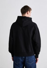 Čierna mikina s kapucňou a voľným strihom, vyrobená z jemnej tkaniny. Má jednoduchý dizajn s rebrovanými manžetami a lemom. Nosí sa s svetlomodrými džínami.