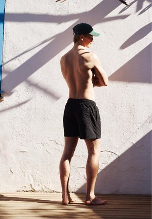 Mężczyzna bez koszulki, w czarnych szortach i zielonej czapce, stoi boso na drewnianej podłodze, zwrócony twarzą do białej ściany z ukośnymi cieniami.