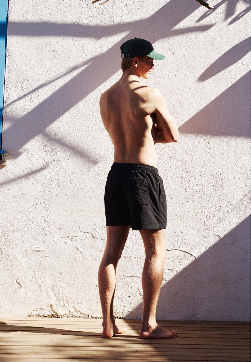 Homme torse nu portant un short noir et une casquette verte, debout pieds nus sur un sol en bois, face à un mur blanc avec des ombres diagonales.