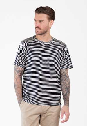 Man with tattooed arms wearing a black and white striped T-shirt and beige pants, looking to his left with hands in pockets.