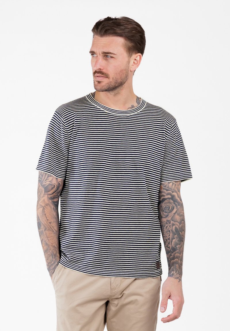 Hombre con brazos tatuados, vistiendo una camiseta de rayas blancas y negras y pantalones beiges, mirando hacia su izquierda con las manos en los bolsillos.