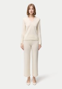 Cremefarbener Strickpullover mit V-Ausschnitt und Spitzenbesatz, kombiniert mit passenden, weit geschnittenen Hosen mit ausgefranstem Saum. Das Model steht vor einem neutralen Hintergrund.