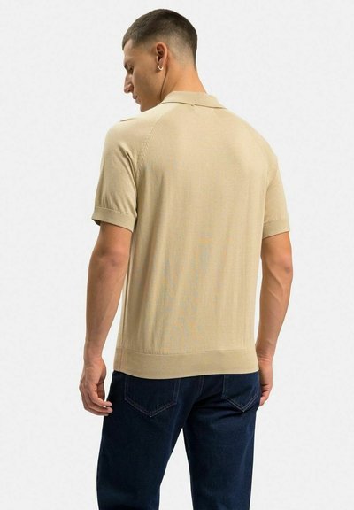 Beige kortärmad polo med ribbad krage och kant, med en slät textur och figurnära passform, bärs tillsammans med mörka denimjeans.